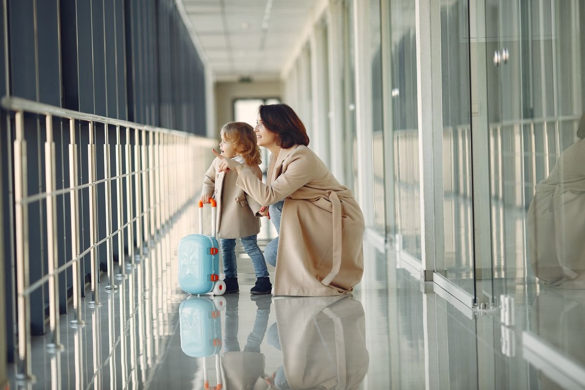 Mother and young child in an airport hallway with a suitcase, looking out at the view